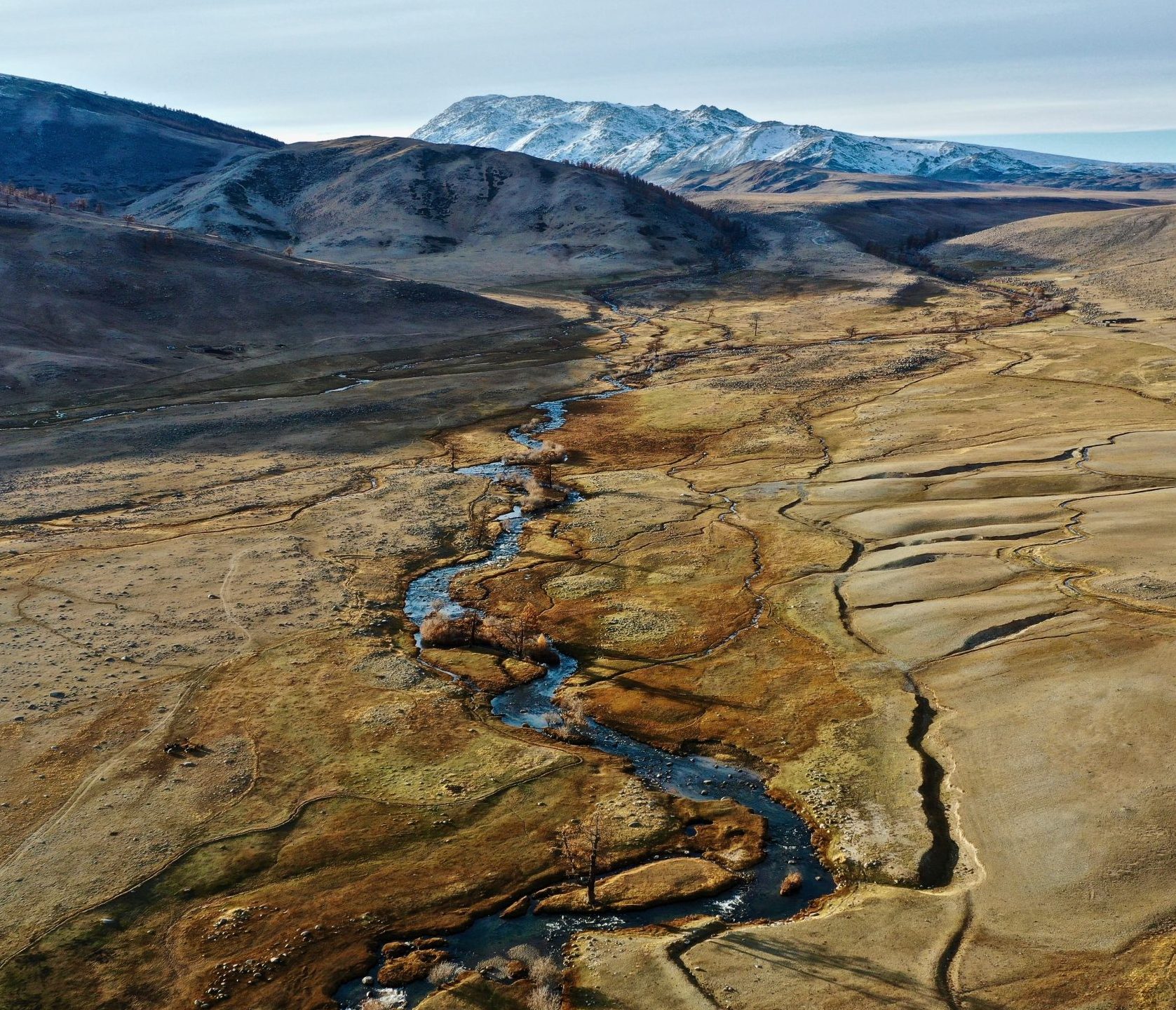 Aerial view of a river winding through dry grassland below snow-topped mountains, a setting for geological gas surveys.